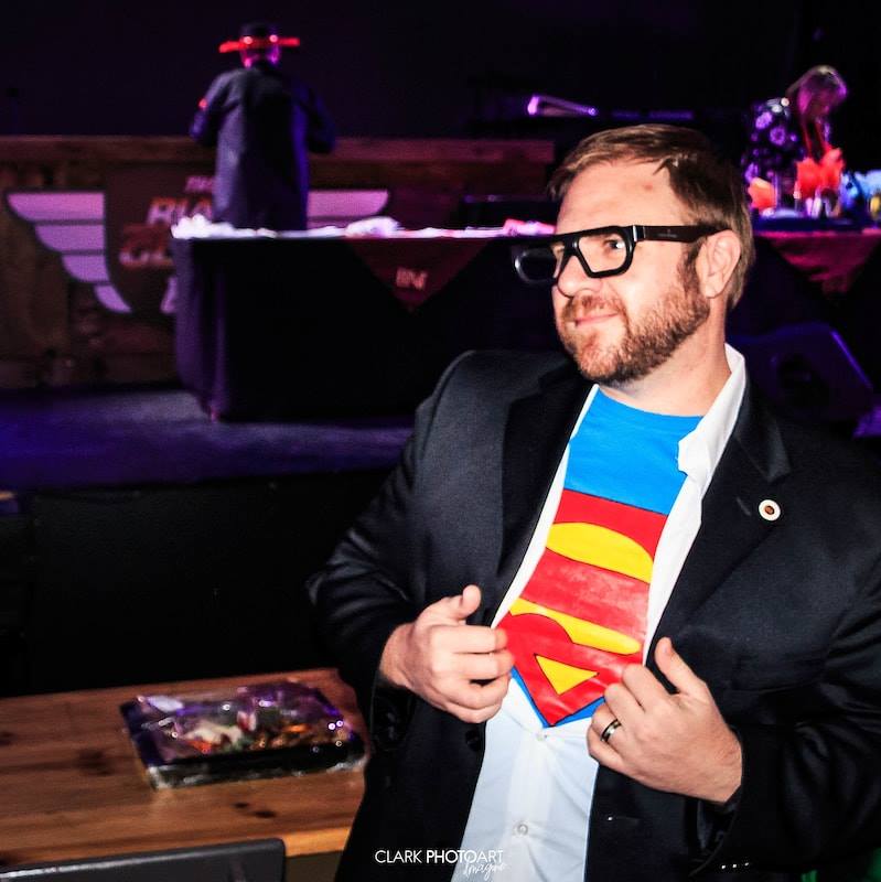 Warren Pridgeon pulling open a suit jacket to reveal a Superman shirt, on stage